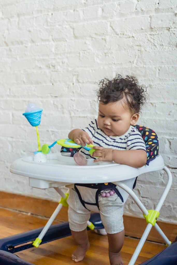 Curious little barefoot African American baby sitting in walker and playing with toy near white wall in room at home